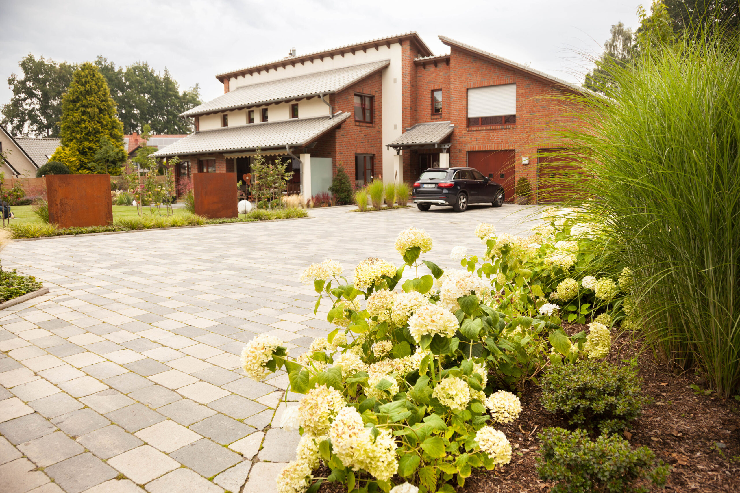 Modernes zweistöckiges Backsteinhaus mit gepflegtem Garten und geparktem Auto auf gepflastertem Hof.