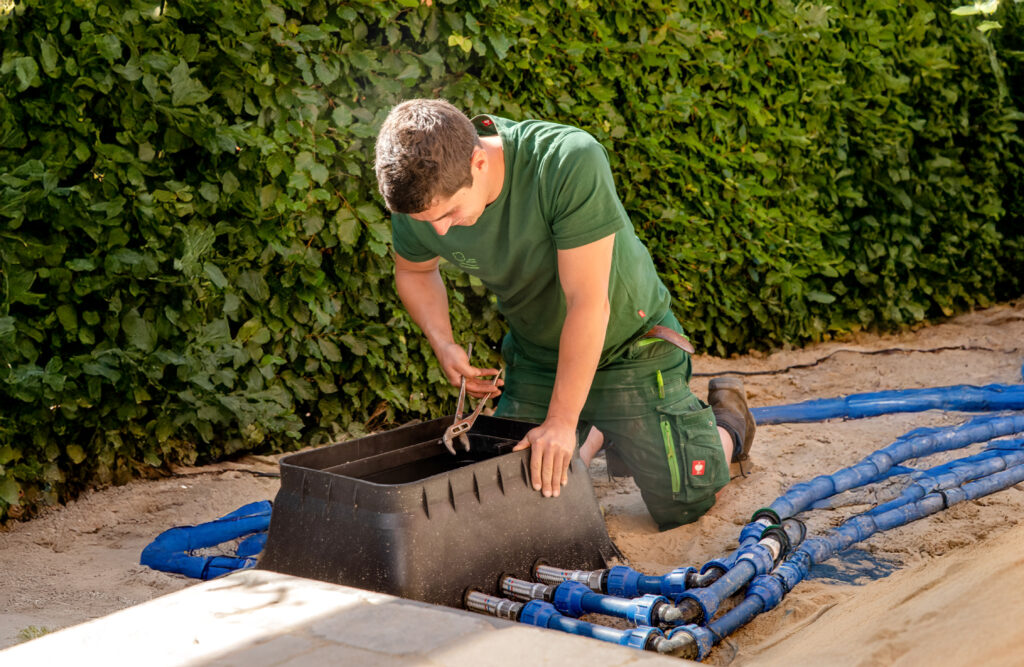 Person in grüner Arbeitskleidung arbeitet mit Werkzeug an schwarzem Kasten, umgeben von blauen Schläuchen.