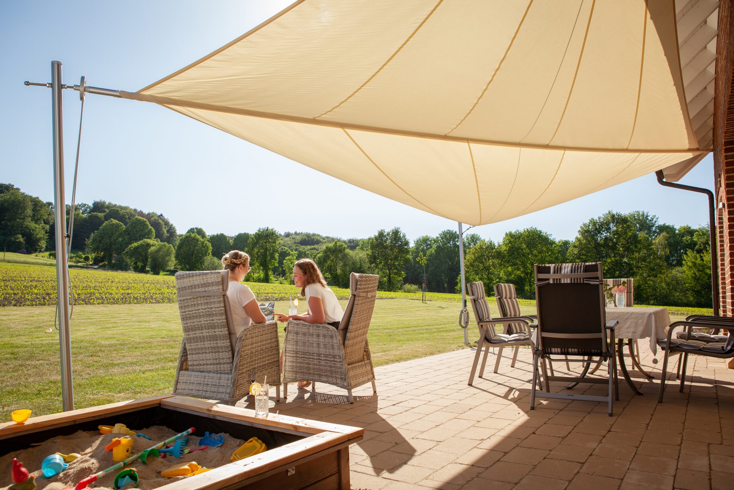 Zwei Personen entspannen sich unter einem Sonnensegel auf einer Terrasse mit Sandkasten und Spielzeug.
