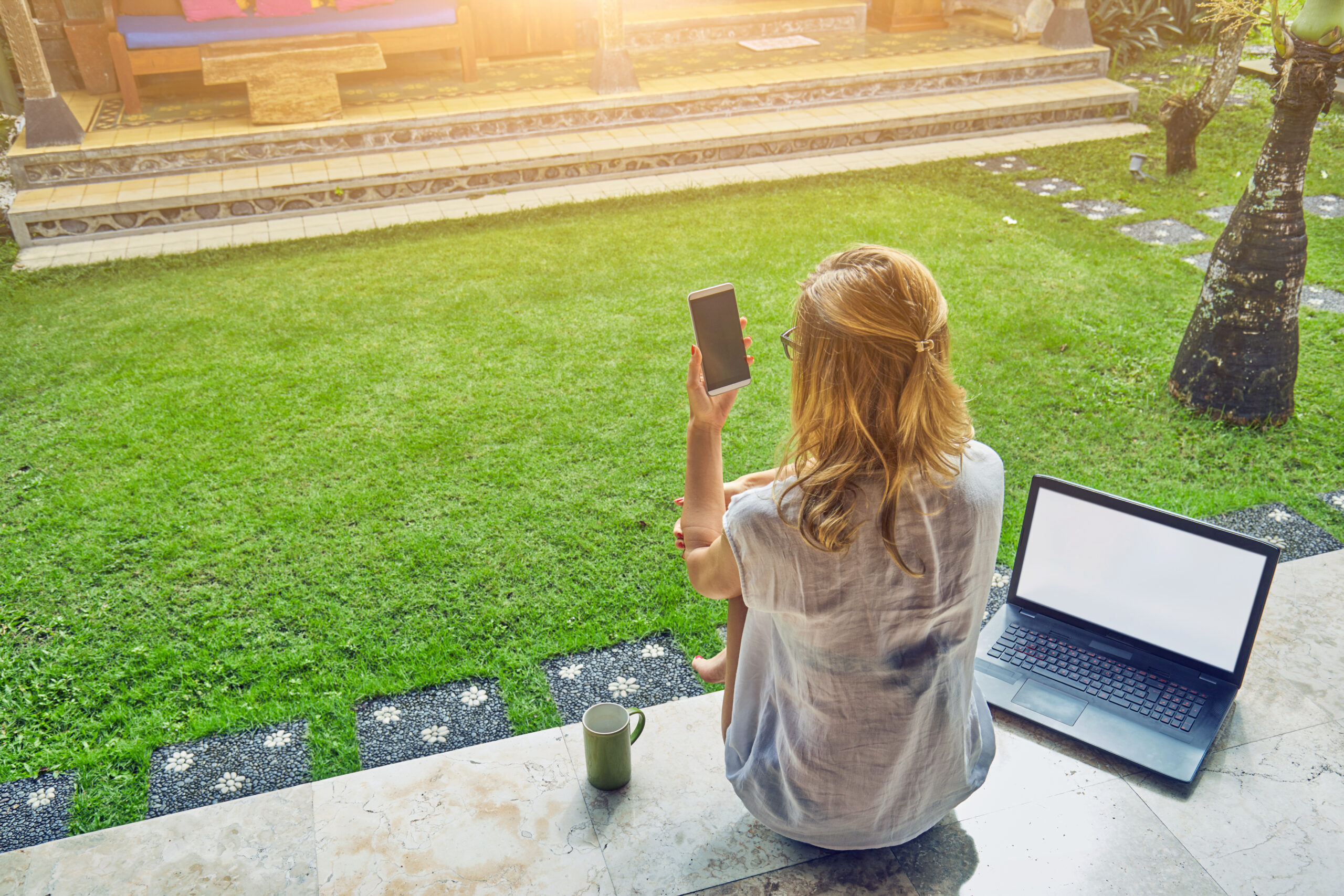 Eine Frau sitzt auf einer Terrasse, vertieft in ihr Smartphone, mit Blick auf einen grünen Garten.