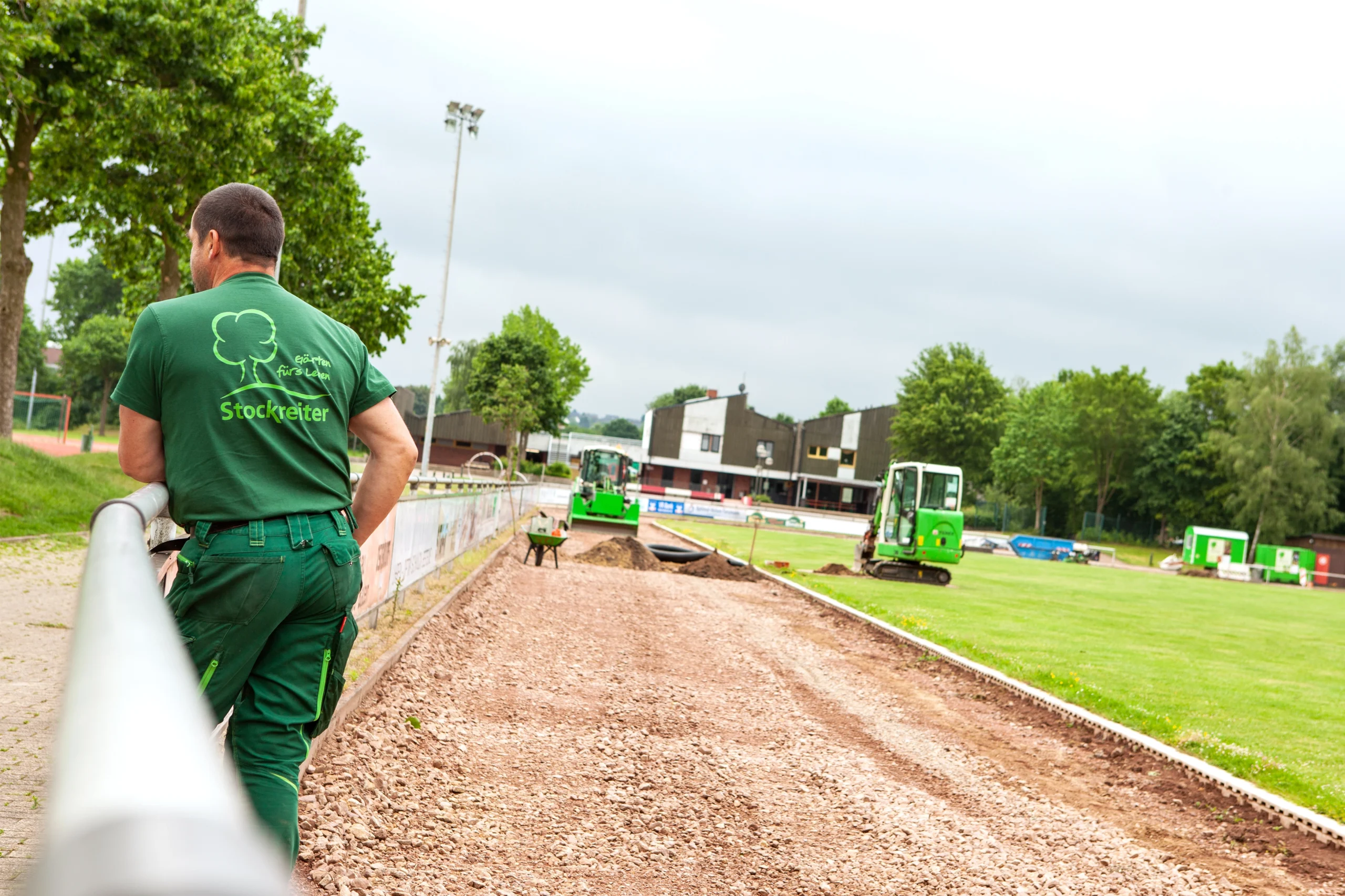 Baustelle auf Sportplatz: Person in grüner Kleidung beobachtet Baggerarbeiten auf erdigem Boden.