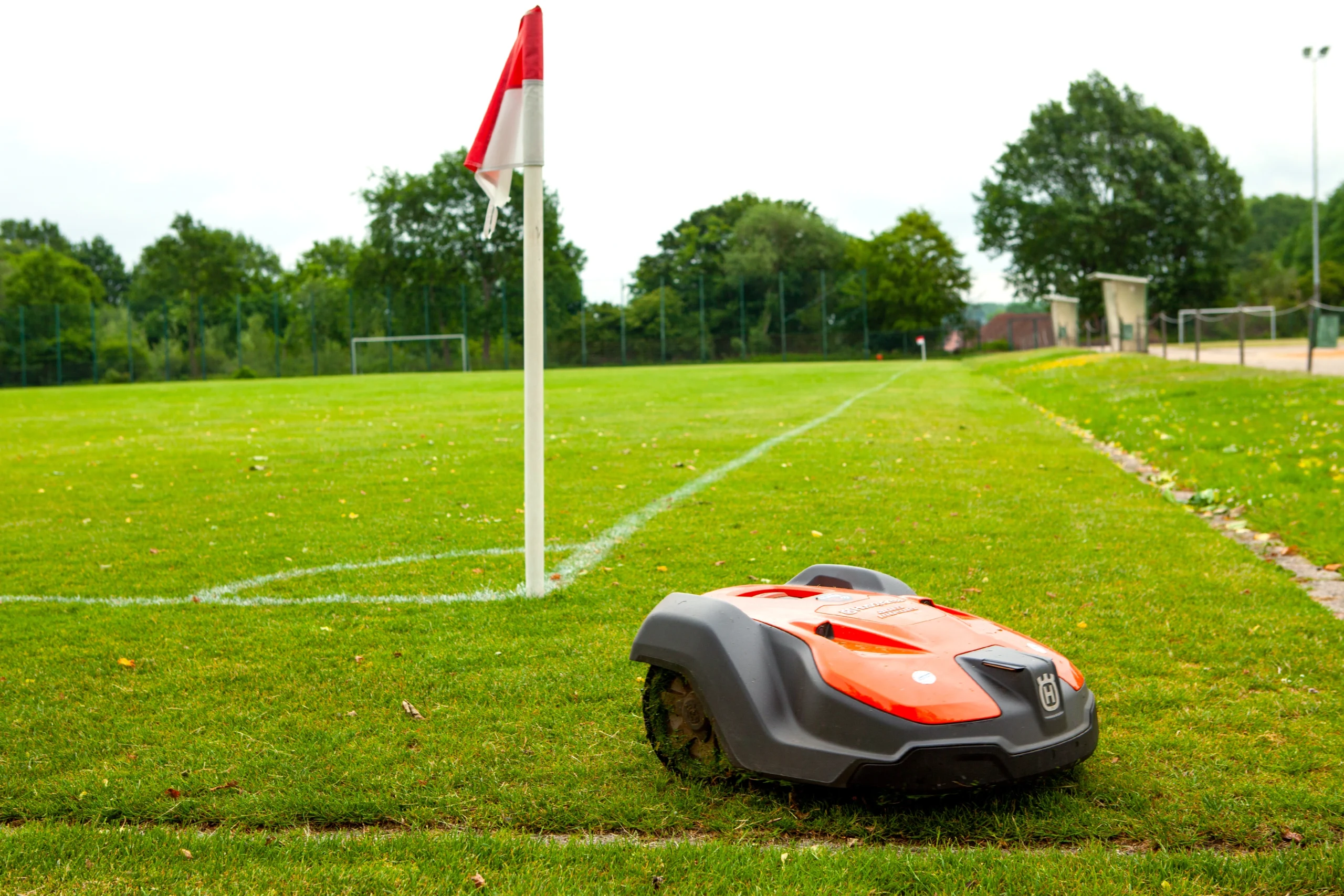 Ein automatischer Rasenmäher mäht den grünen Rasen eines Sportplatzes neben einer rot-weißen Eckfahne.