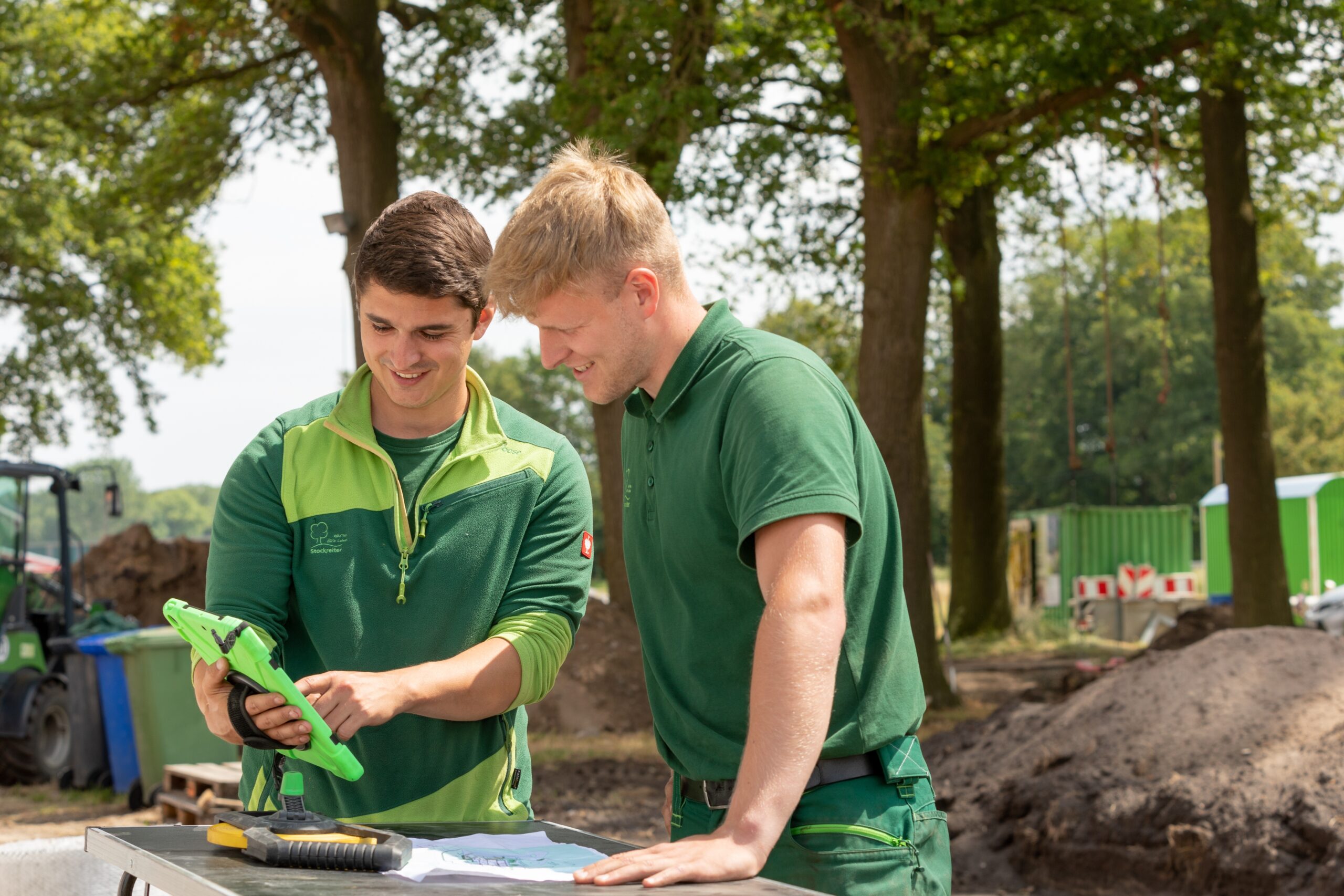 Zwei Männer in grüner Arbeitskleidung betrachten ein Tablet auf einer Baustelle im Freien.