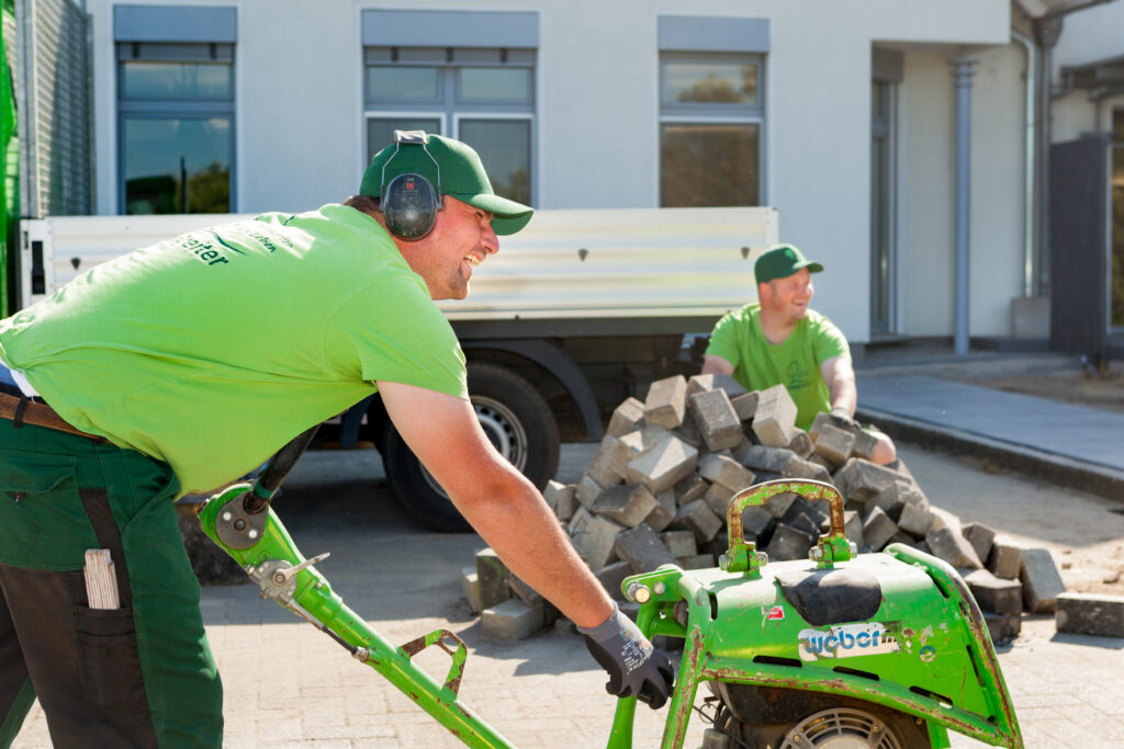 Zwei Arbeiter in grüner Kleidung arbeiten auf einer Baustelle mit Pflastersteinen und Baumaschine.