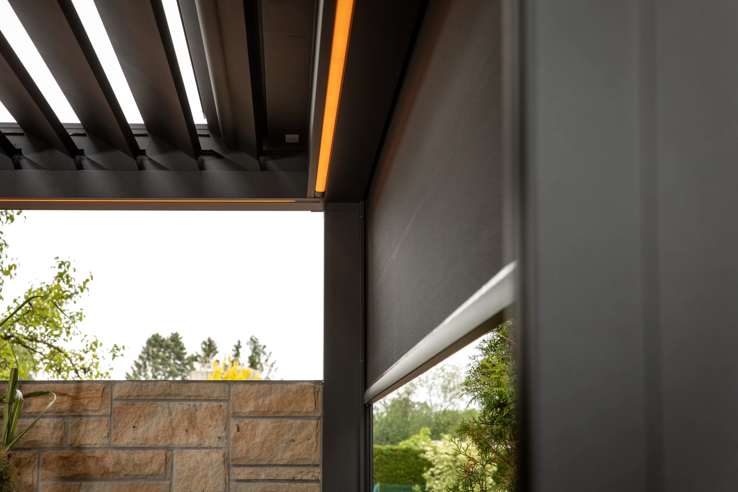 Moderne Pergola mit schrägen Lamellen, dezenter Beleuchtung und Steinmauer in einem eleganten Garten.