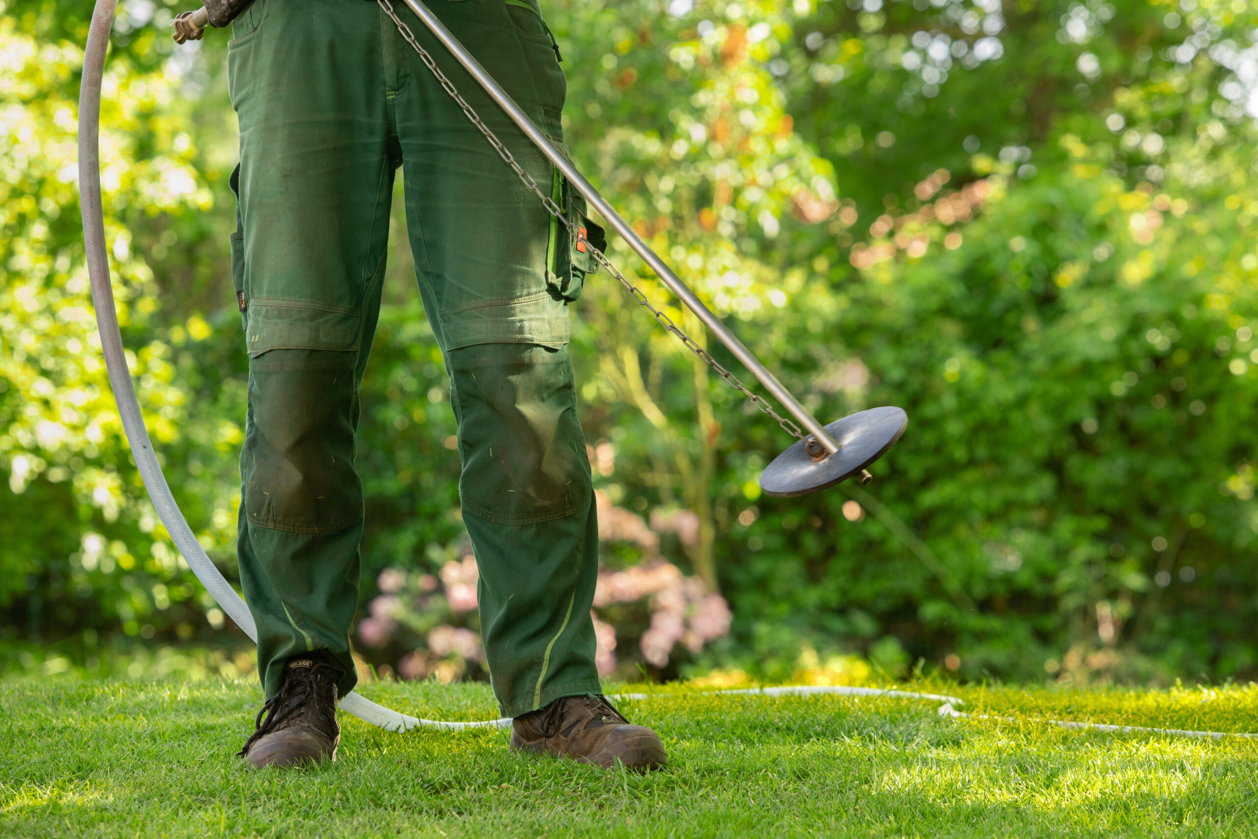 Person in grüner Arbeitskleidung bearbeitet mit einem Bodenwerkzeug den Rasen auf einer Grünfläche.