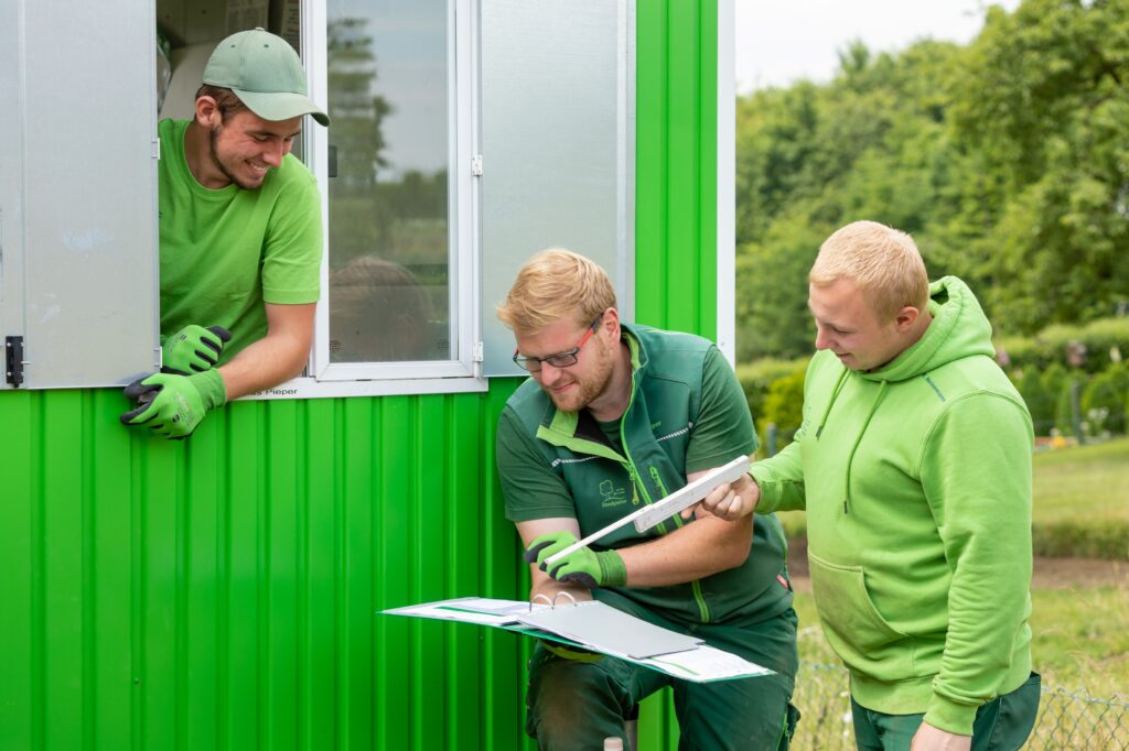 Drei Männer in grüner Arbeitskleidung besprechen Dokumente vor einem grünen Gebäude im Freien.