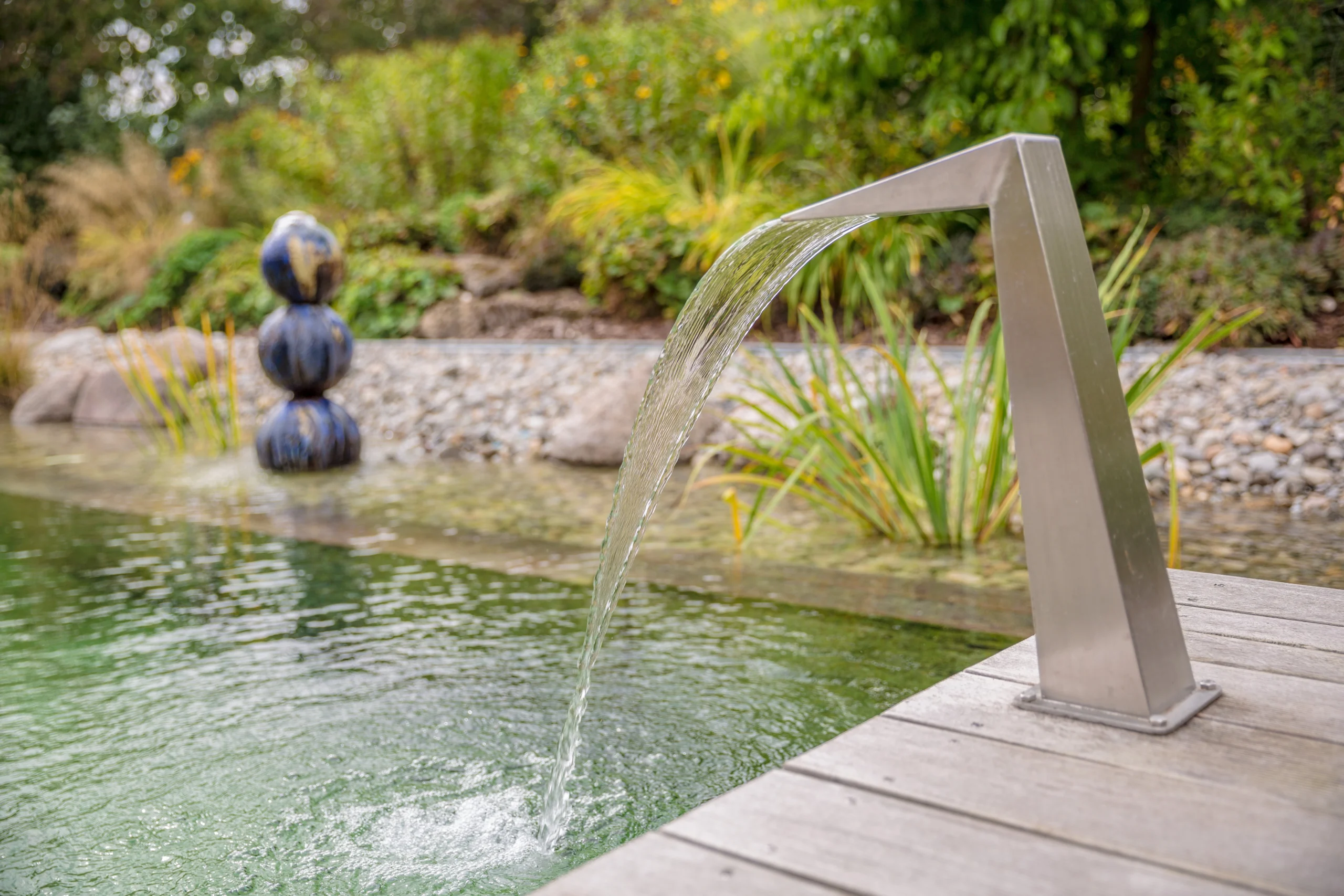 Modernes Wasserspiel im Garten: Wasser fließt aus einem Rohr in einen Teich, umgeben von Pflanzen.