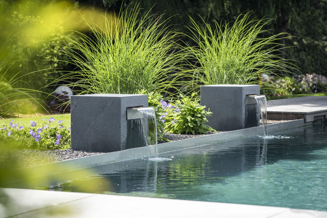 Moderne Gartenszene mit fließendem Wasser aus grauen Steinstrukturen, umgeben von grünen Pflanzen.