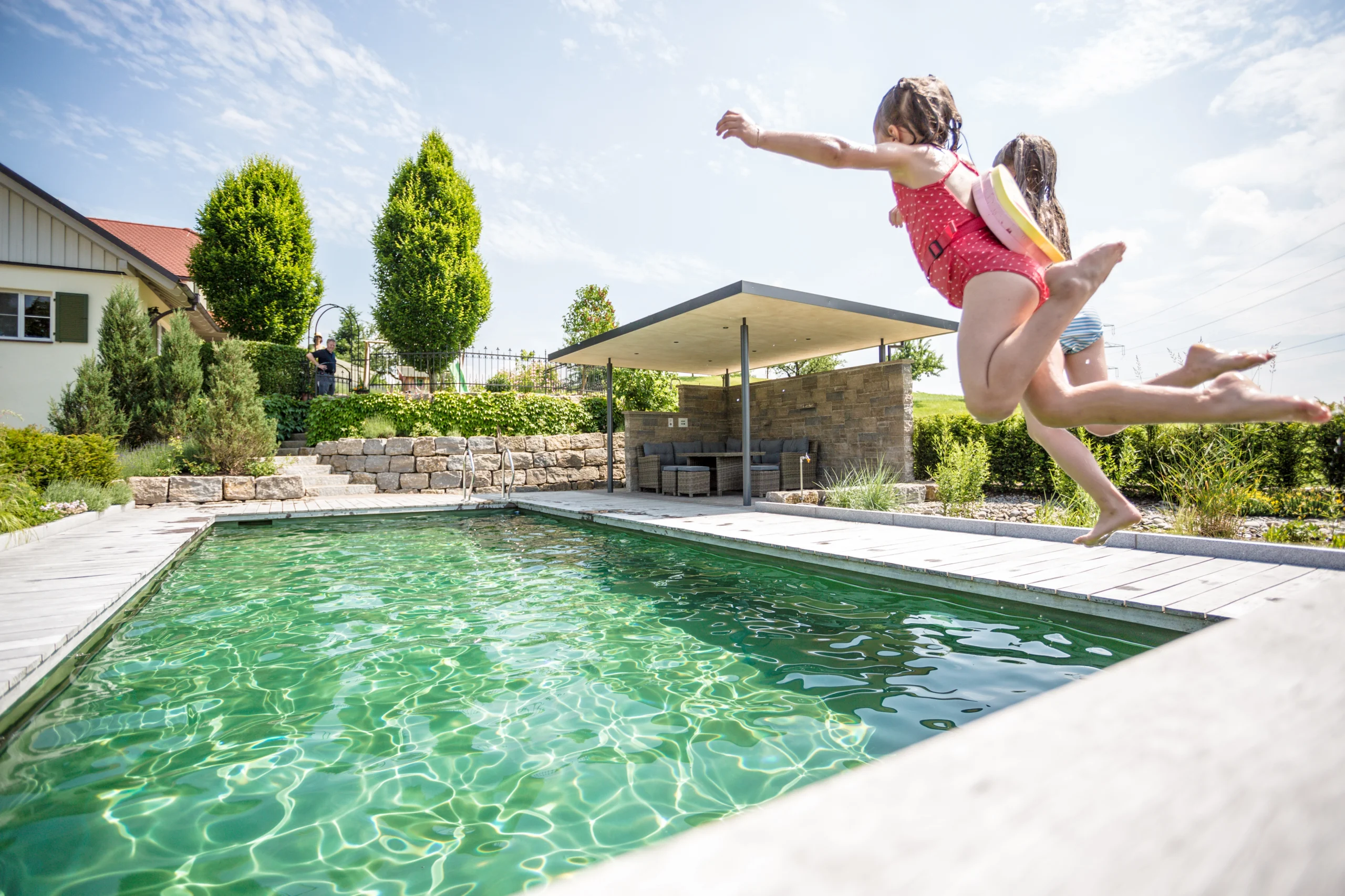 Zwei Kinder springen fröhlich in ein grünes Schwimmbecken im sonnigen Garten mit Sitzbereich.