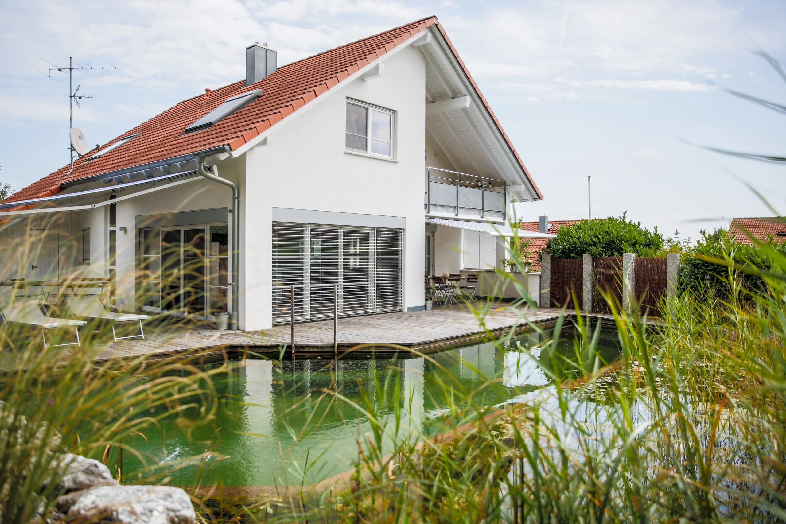 Modernes Haus mit rotem Dach, weißer Fassade, großem Balkon und Naturteich mit Holzdeck und Pflanzen.
