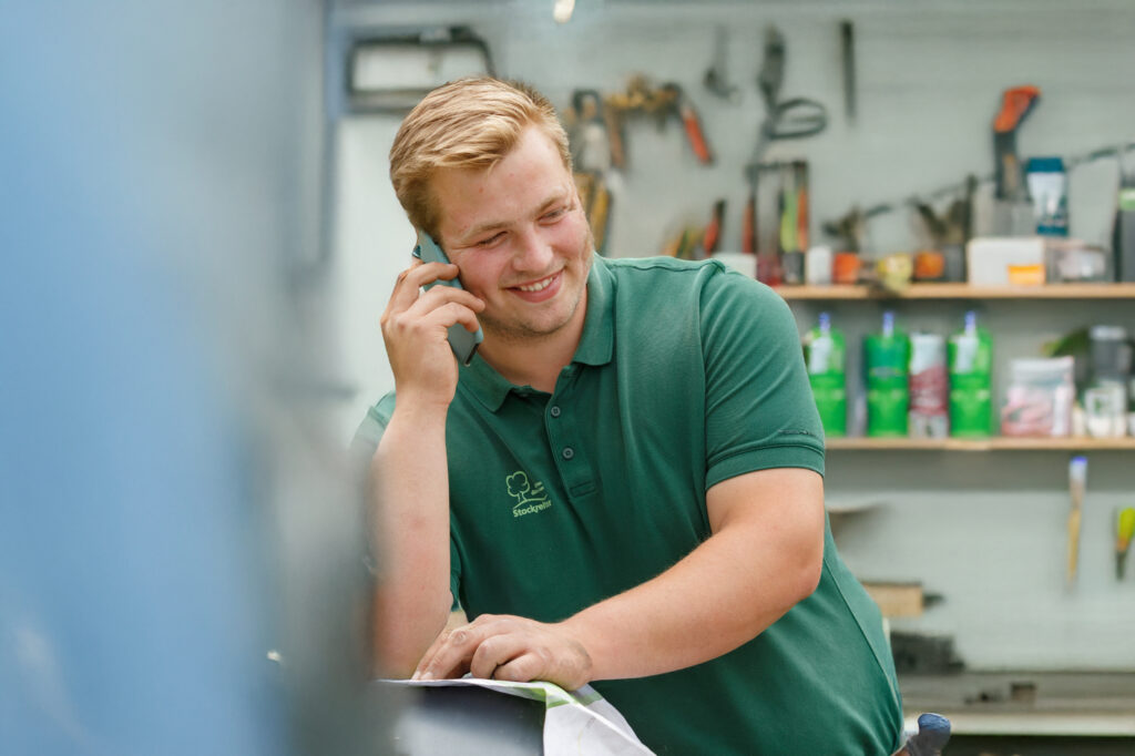 Ein junger Mann in grünem Poloshirt telefoniert lächelnd in einer Werkstatt mit Werkzeugen im Hintergrund.