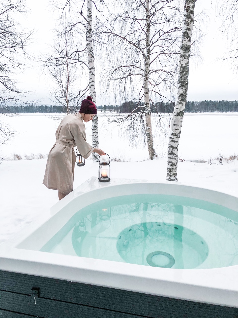 Person im Bademantel mit Laterne neben einem Whirlpool im Schnee, umgeben von schneebedeckten Bäumen.