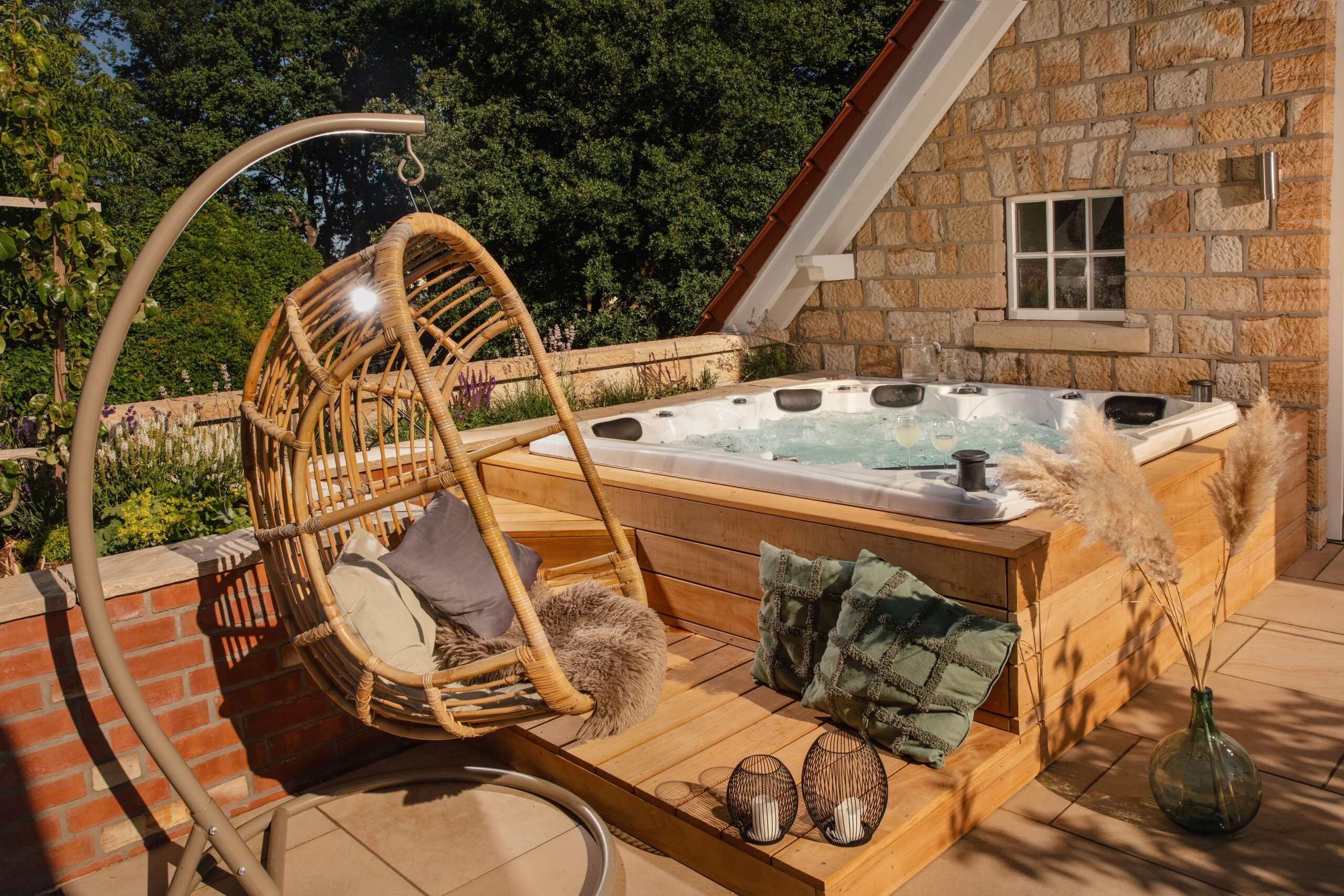 Gemütliche Terrasse mit Whirlpool, Getränken, Korbsessel und rustikaler Steinmauer, umgeben von Grün.