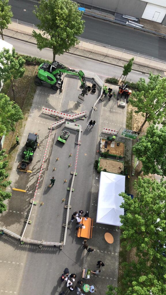 Vogelperspektive auf eine Messe im Freien mit grünen Baggern, Zelt und Menschen in Gesprächen.