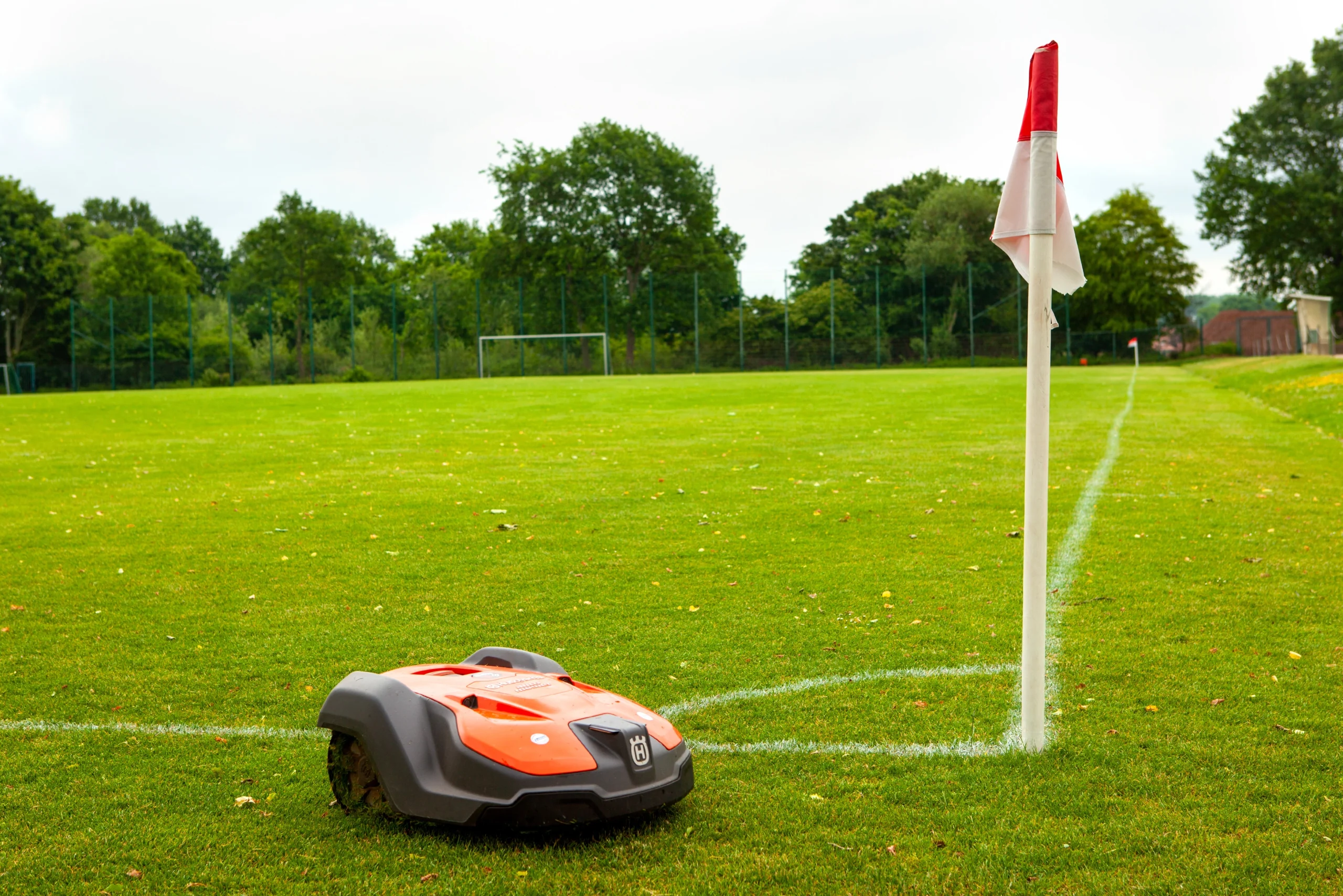 Ein orangefarbener automatischer Rasenmäher pflegt den grünen Sportplatz mit Tor und Eckfahne.