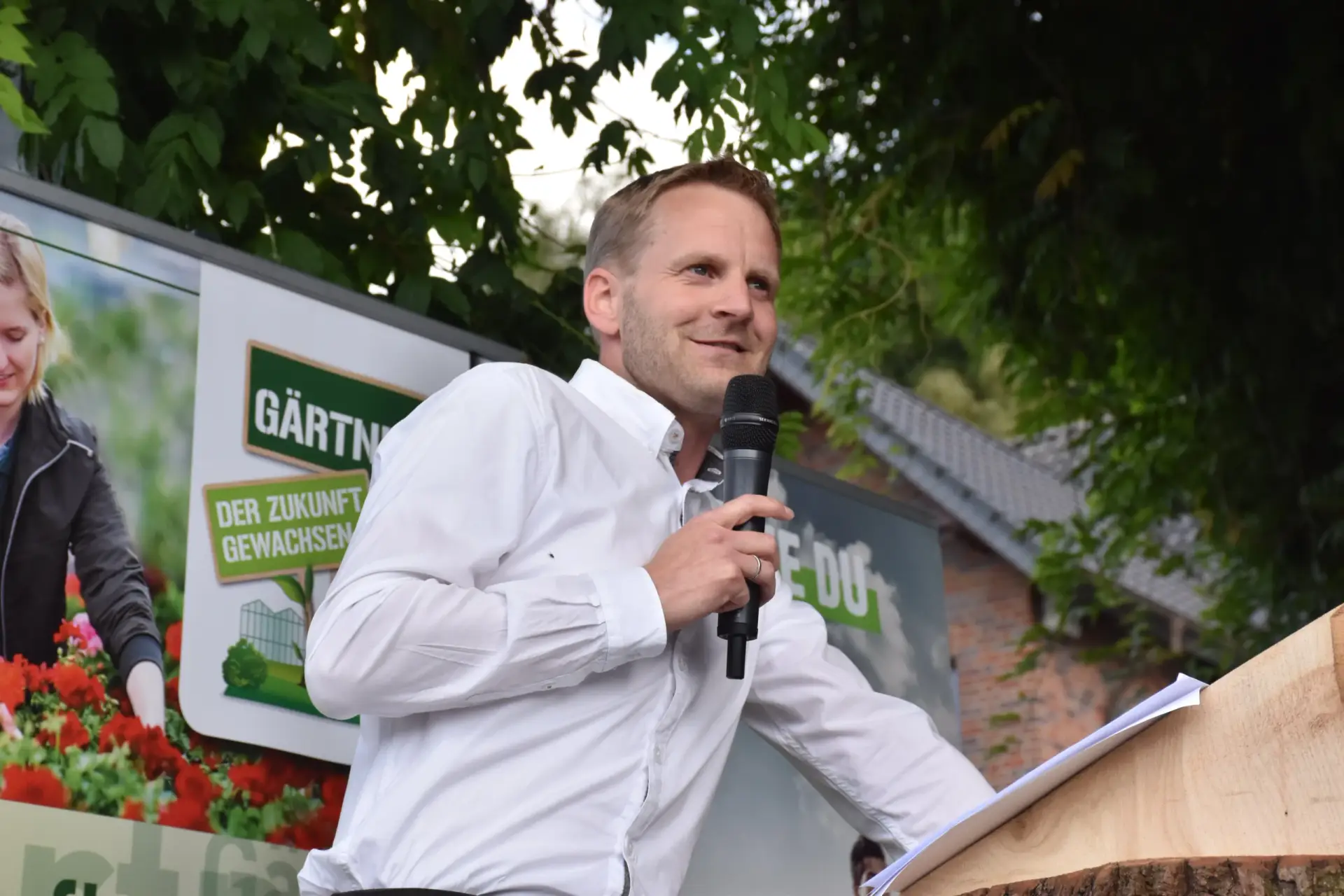 Ein Mann im weißen Hemd hält ein Mikrofon vor einem Plakat mit "GÄRTNEREI DER ZUKUNFT GEWACHSEN".
