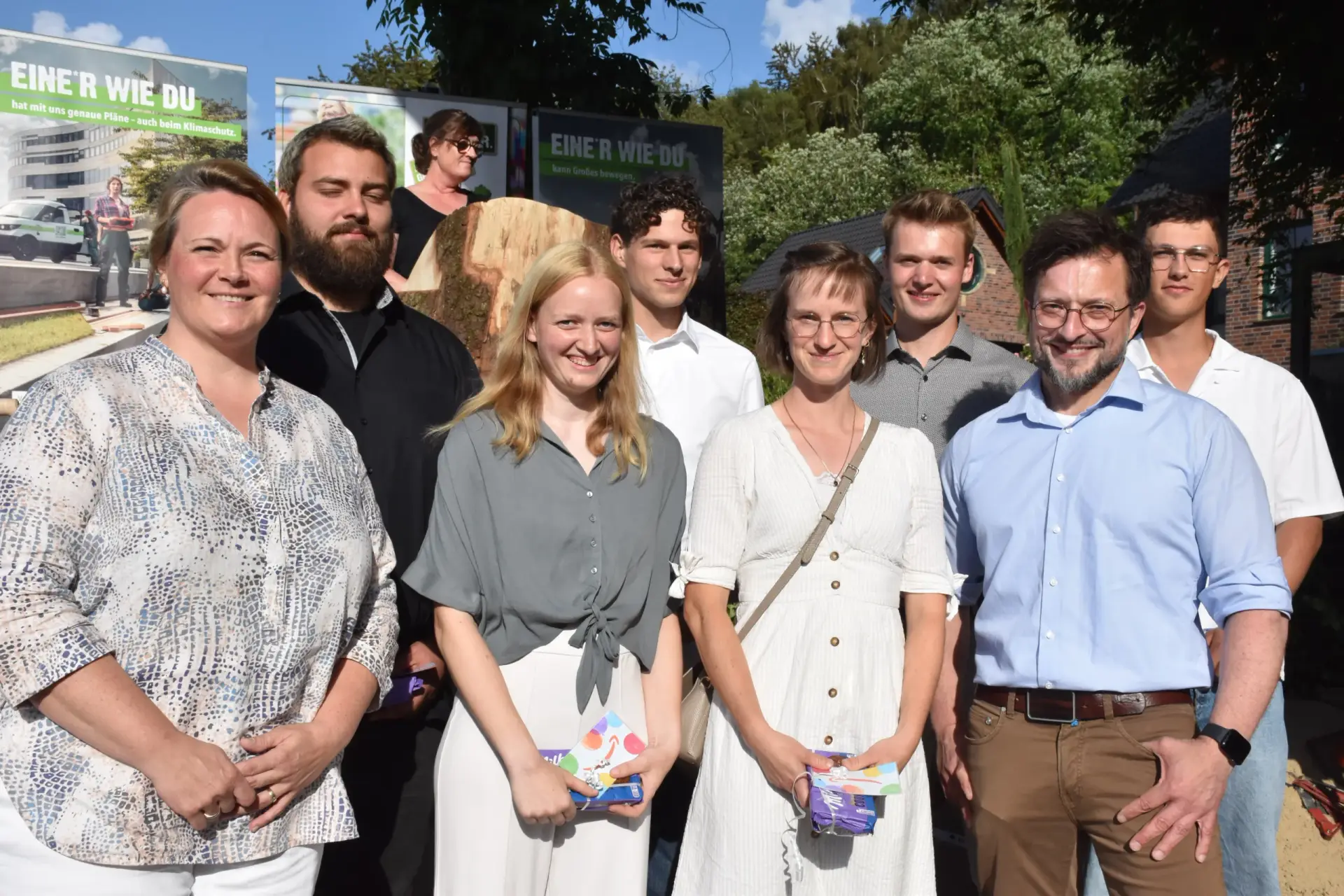 Eine Gruppe von acht lächelnden Personen posiert im Freien vor Plakaten mit der Aufschrift „EINE*R WIE DU“.
