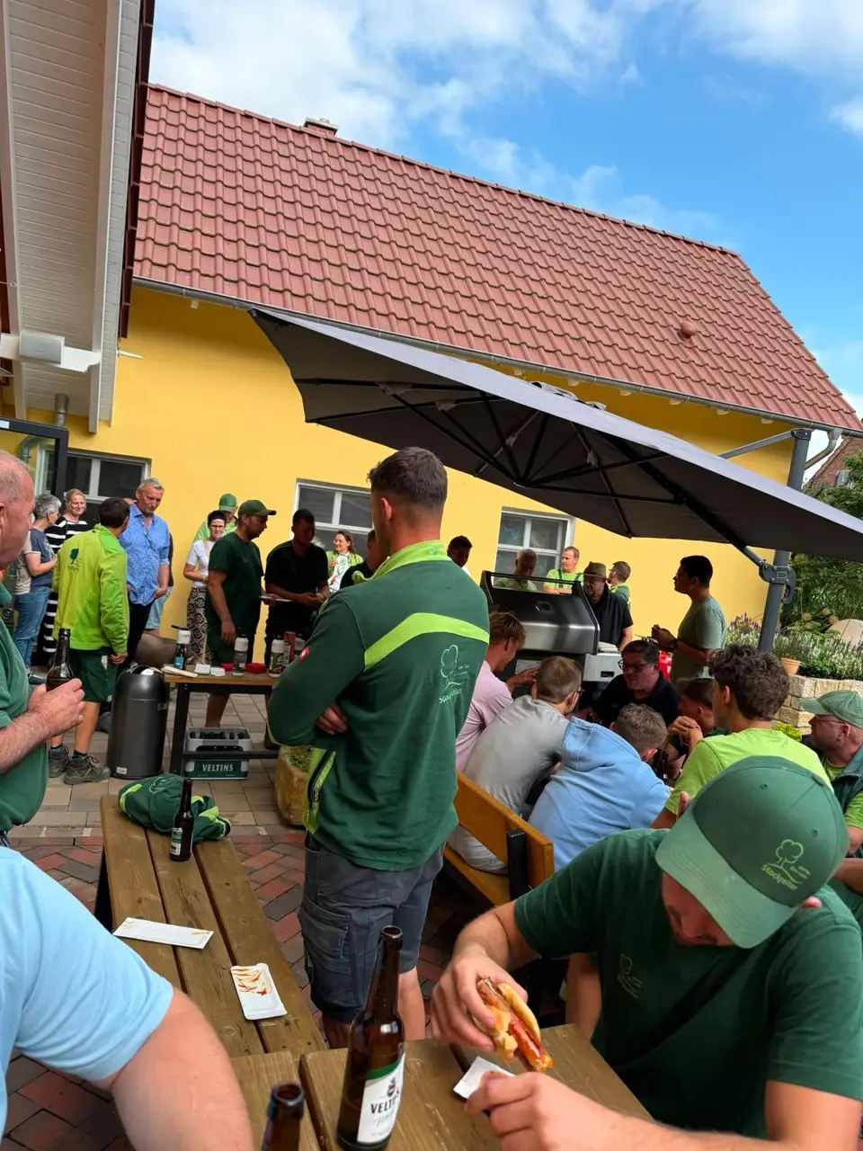 Menschen bei einem fröhlichen Grillfest im Freien, umgeben von Grün und einem gelben Gebäude im Hintergrund.