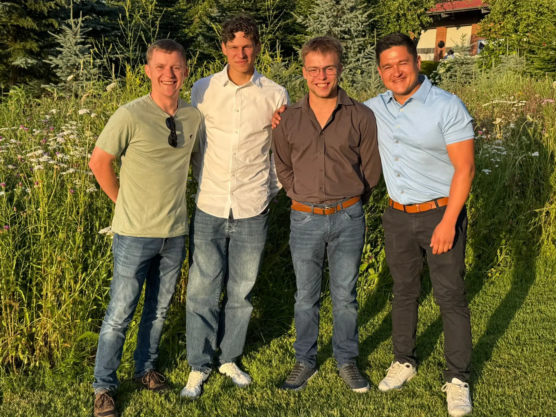 Vier lächelnde Männer in Freizeitkleidung stehen eng beieinander vor üppiger, grüner Vegetation.