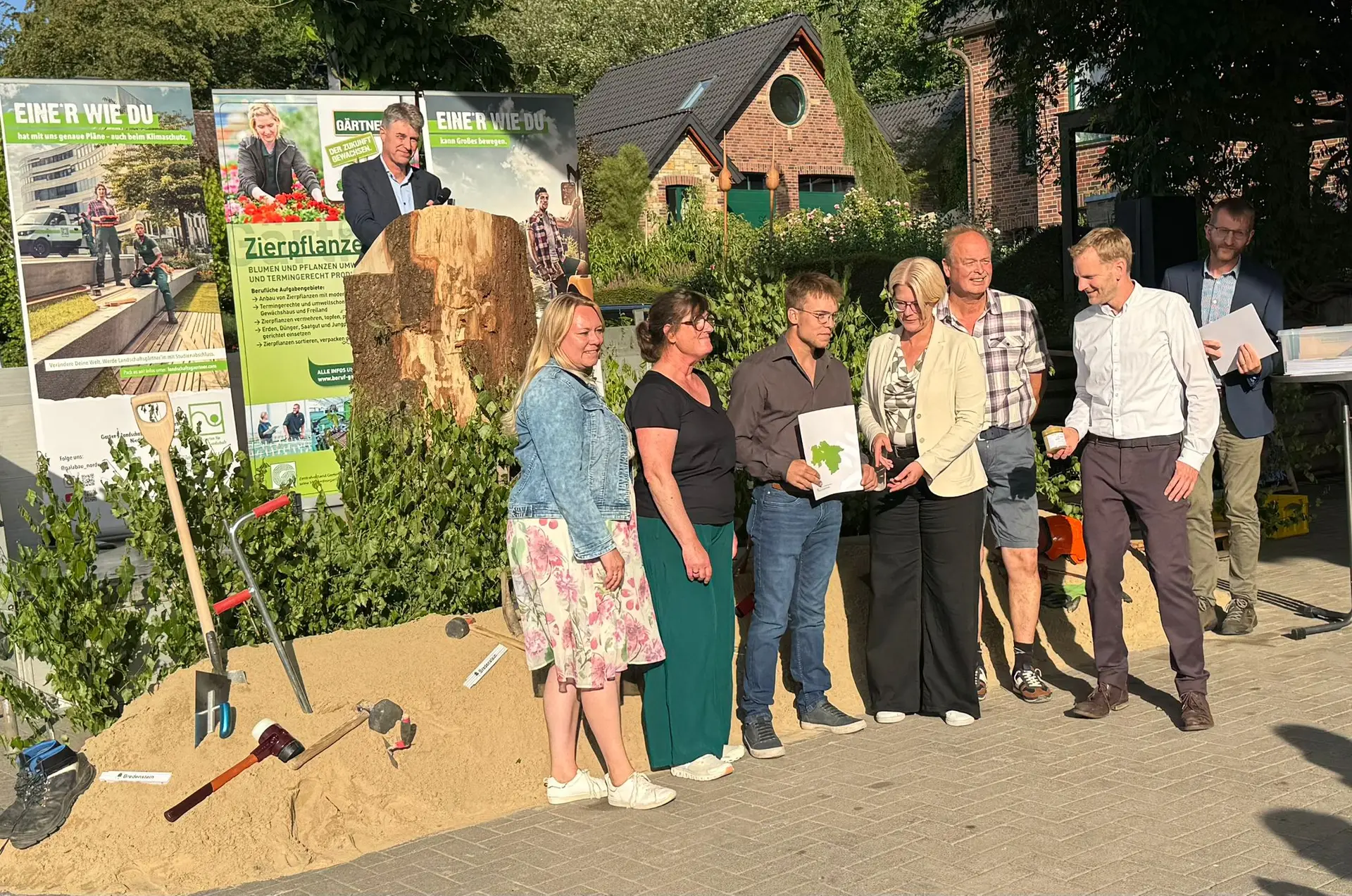 Eine Gruppe von sieben Personen posiert lächelnd vor Plakaten zu Gartenarbeit und Klimaschutz.