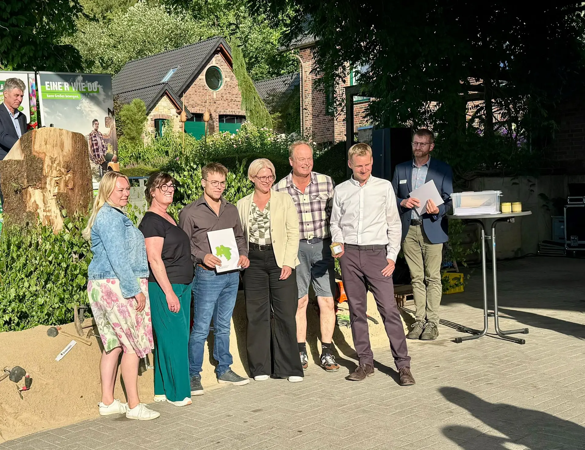 Sieben Personen posieren draußen, eine hält ein Zertifikat. Im Hintergrund ein Plakat: „EINER WIE DU...“.