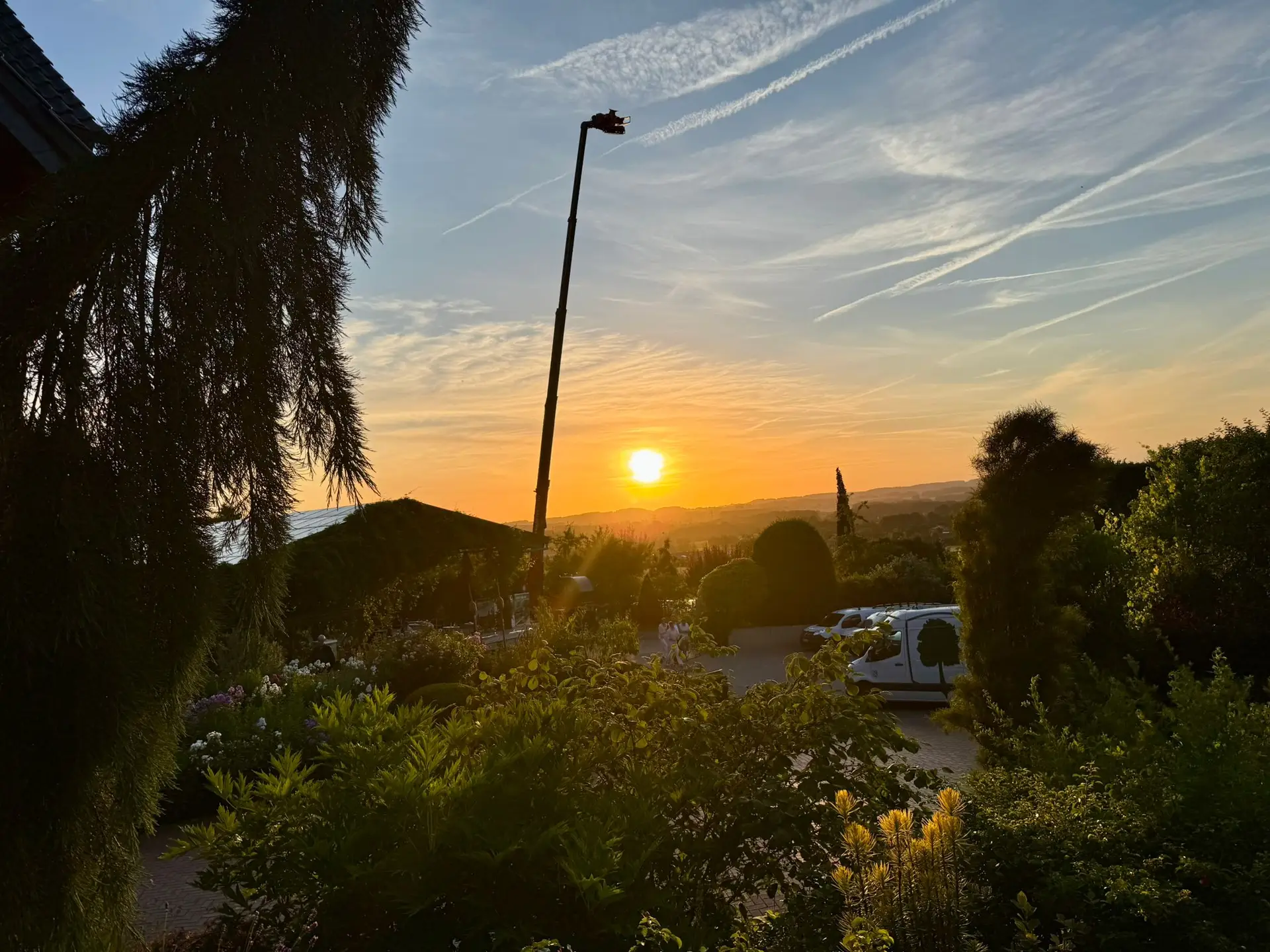 Sonnenuntergang über üppiger Landschaft mit Baum, geparktem Lieferwagen und Gebäude am Rand.