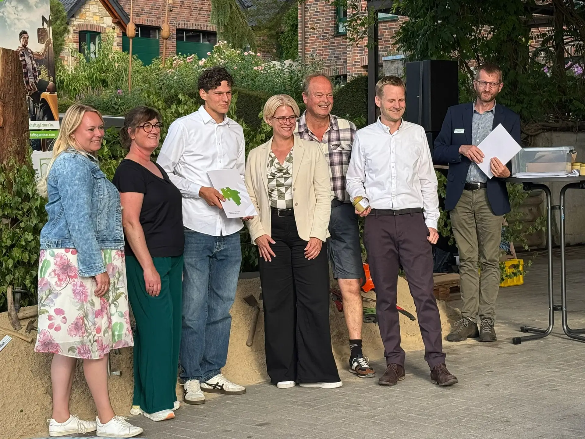 Eine Gruppe von sieben Personen posiert lächelnd vor einem Backsteingebäude mit Pflanzen im Hintergrund.
