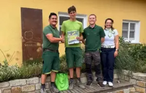 Vier Personen vor gelbem Gebäude, zwei in Arbeitskleidung, schütteln Hände, Buch "Gartengestaltung".