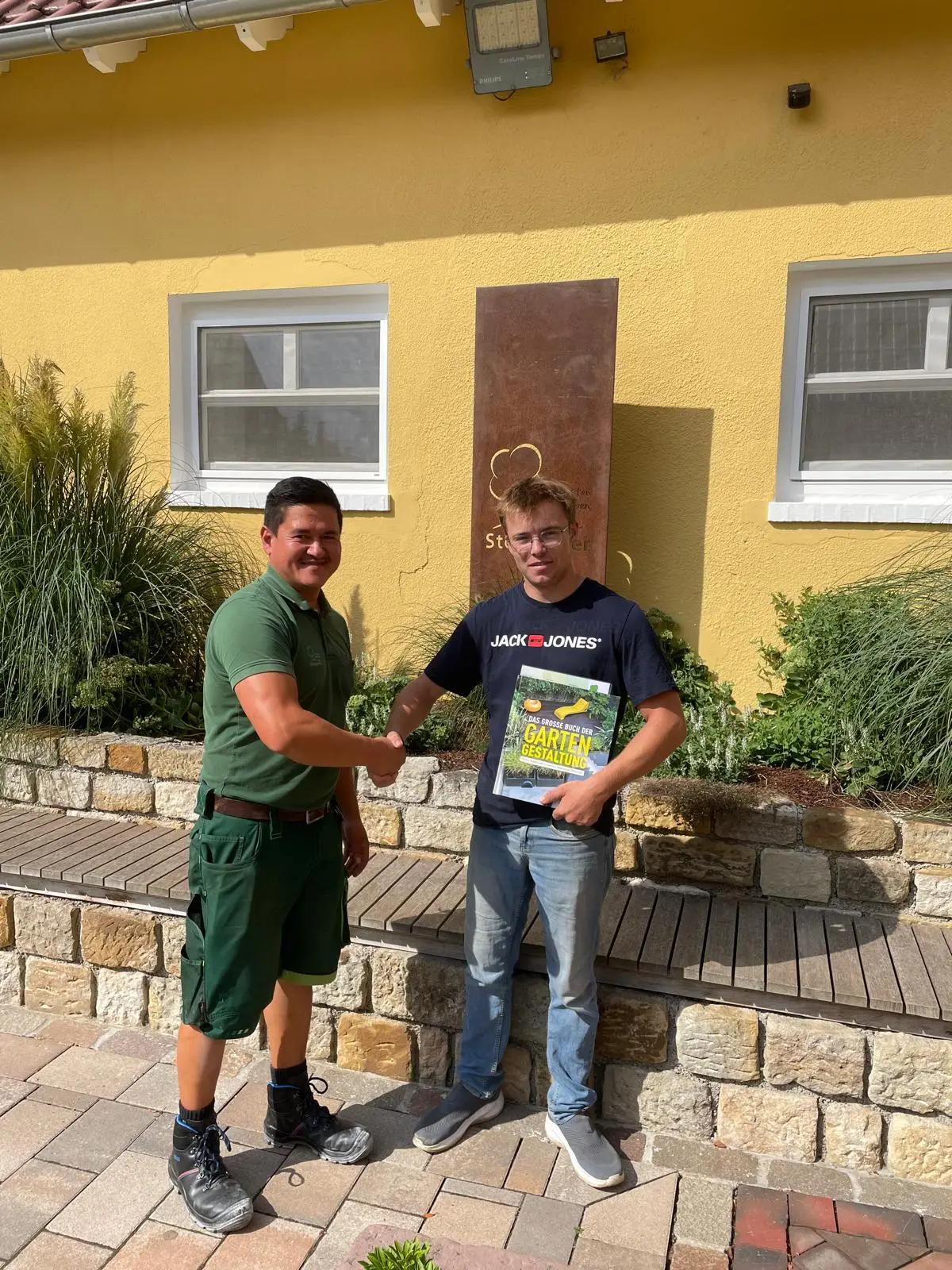 Interne Lossprechung und Ausbildung bei Stockreiter 6 Zwei Personen geben sich die Hand vor einer gelben Wand, eine hält ein Buch über Gartengestaltung.