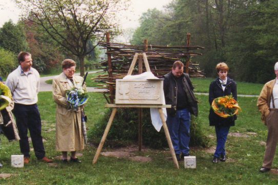 Sechs Personen stehen bei einer Gedenkveranstaltung im Freien, halten Blumen und eine Tafel ist sichtbar.