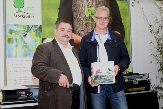 Zwei Männer präsentieren ein Buch; einer hält ein Mikrofon. Im Hintergrund ein Plakat zu Gartenpflege.