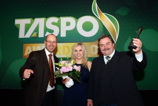 Drei Personen posieren lächelnd vor dem "TASPO AWARD"-Hintergrund; eine hält eine Trophäe hoch.