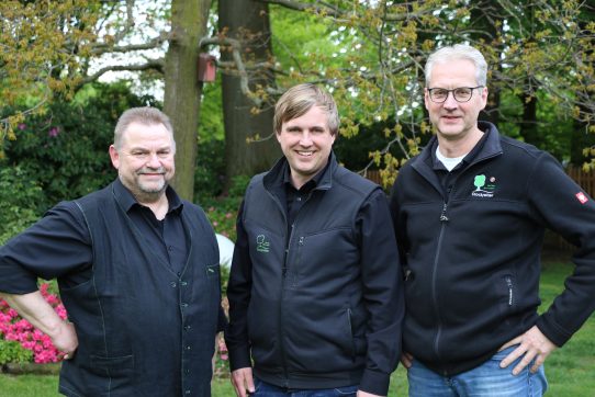 Drei lächelnde Männer in dunkler Kleidung stehen entspannt in einem grünen Garten mit rosa Blüten.