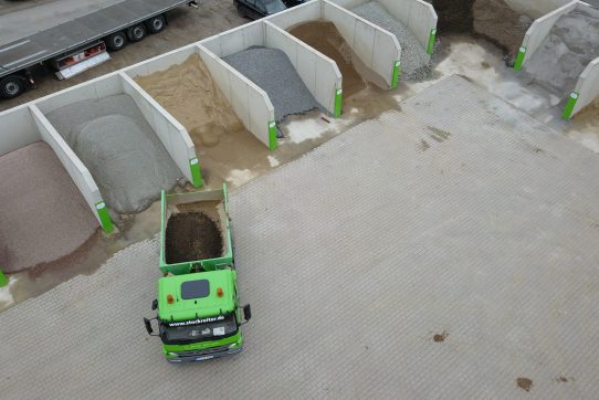 Ein grüner Lastwagen steht auf einem Lagerplatz mit Betonboxen voller Sand, Kies und Erde.