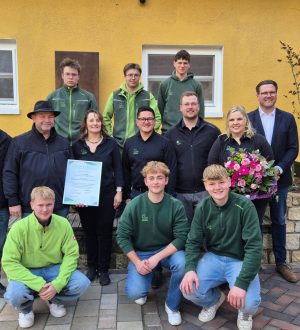 Eine Gruppe von 13 Personen posiert lächelnd vor einem gelben Gebäude, eine Person hält eine Urkunde.