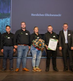 Sieben Personen posieren lächelnd bei einer Preisverleihung. Eine Person hält Blumen, eine andere eine Urkunde.