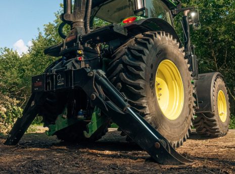 Ein großer grüner Traktor steht still auf einem erdigen Feld, umgeben von Bäumen unter blauem Himmel.