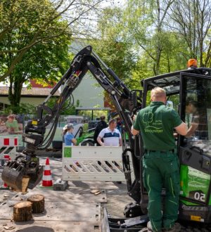 Ein Arbeiter bedient eine grüne Baumaschine neben Baumstämmen auf einer städtischen Baustelle.