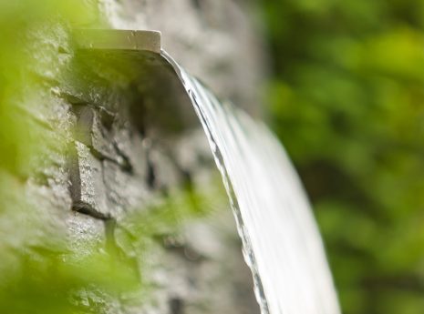 Ein kleiner Wasserfall fließt über eine Metallkante vor einer Steinmauer, umgeben von grünen Pflanzen.