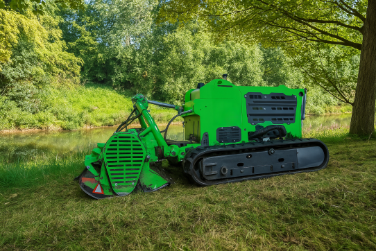 Ein leuchtend grüner Mulcher steht still am Ufer eines Flusses, umgeben von grüner Vegetation.