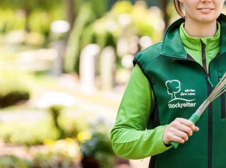 Person in grüner Arbeitskleidung mit Gartengerät, umgeben von Natur, symbolisiert Gartenpflege.