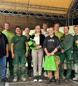 Eine Gruppe von zehn Personen in grüner Arbeitskleidung posiert lächelnd mit "Stockreiter"-Taschen auf einer Bühne.