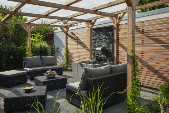 Moderne Terrasse mit Rattanmöbeln, Holzpergola, Wasserbrunnen und üppigen Pflanzen.