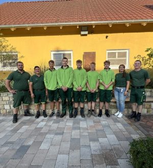 Neun Menschen in grüner Kleidung posieren lächelnd vor einem gelben Gebäude mit rotem Dach.