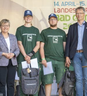 Vier Personen posieren auf einer Bühne; zwei in grüner Arbeitskleidung, zwei formell, mit Trophäen im Hintergrund.