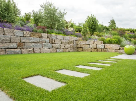 Ein gepflegter Garten mit Rasen, Steinweg, Trockenmauer und bunten Pflanzen unter bewölktem Himmel.