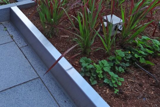 Ein gepflegtes Beet mit roten und grünen Pflanzen, eingefasst von einem silbernen Metallrahmen.