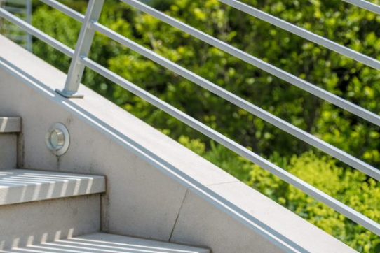 Moderne Außentreppe aus hellem Beton mit metallischem Geländer, umgeben von grünen Bäumen.