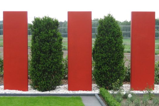 Moderne Gartenlandschaft mit roten Säulen, grünen Büschen und weißen Kieselsteinen auf dem Boden.